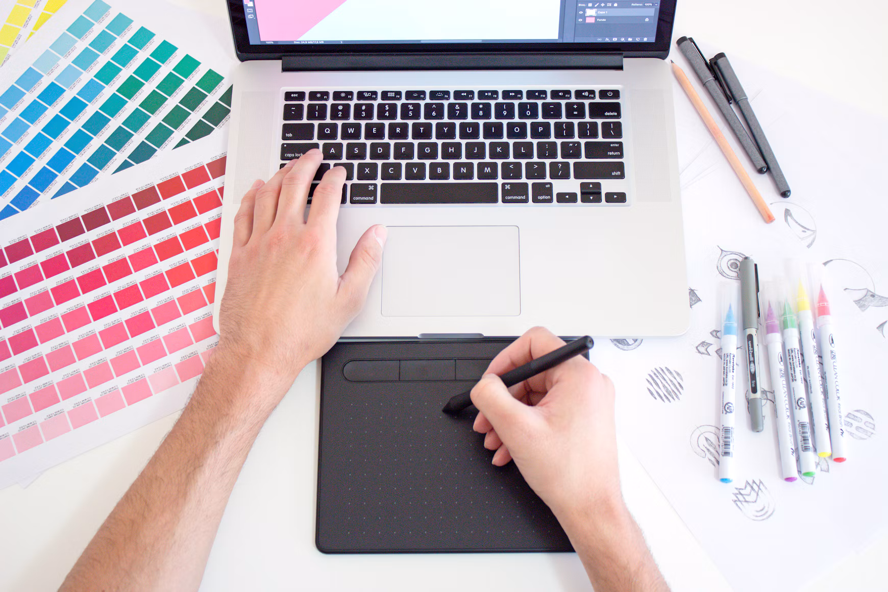 Diseñador trabajando con una tableta gráfica junto a un ordenador, muestras de color y herramientas de dibujo